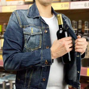 Person concealing a bottle of wine inside their denim jacket in a store symbolizing shoplifting